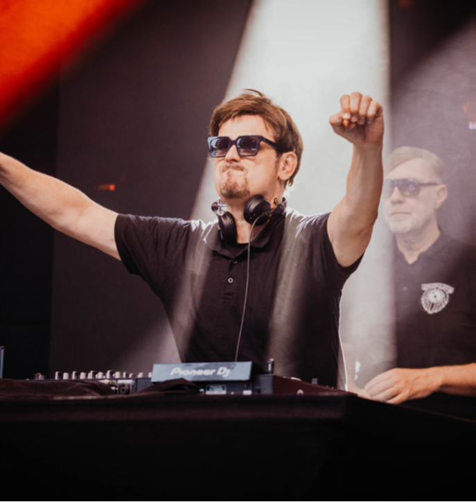DJ mit Kopfhörern hebt die Arme über einem Pioneer DJ-Mischpult auf einer Bühne mit Bühnenlicht.