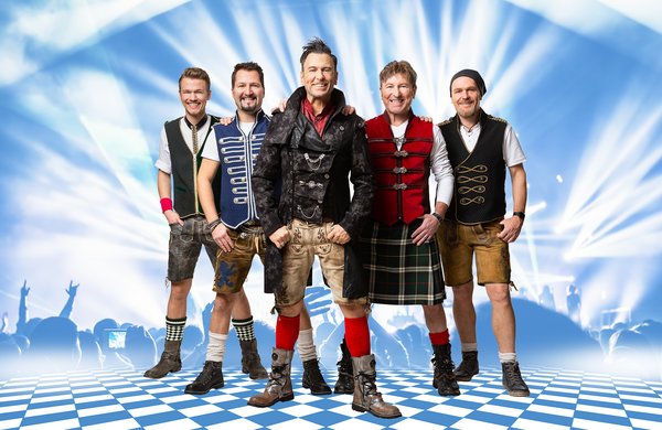 Königlich Bayrisches Vollgas Orchester Fünf Männer stehen auf einem blau-weiß karierten Boden vor einer beleuchteten Bühne. Sie tragen moderne Trachtenmode, darunter Lederhosen, Westen und Stiefel. Der Hintergrund zeigt Lichteffekte und die Silhouetten eines Publikums. Alle lächeln in die Kamera.