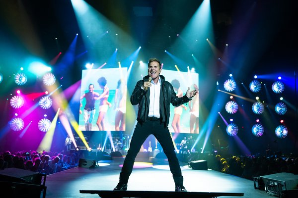 Dieter Bohlen und Band Ein Sänger steht auf einer Bühne bei einem Konzert in einer Arena. Er trägt eine schwarze Lederjacke über einem weißen Hemd und hält ein Mikrofon in der Hand, während er leidenschaftlich singt. Hinter ihm leuchtet ein großer Bildschirm mit einem bunten Bild von tanzenden Menschen am Strand. Die Bühne ist in farbenfrohes Licht getaucht, mit zahlreichen Scheinwerfern, die das Publikum in verschiedenen Farben anstrahlen. Die Stimmung im Publikum wirkt ausgelassen.