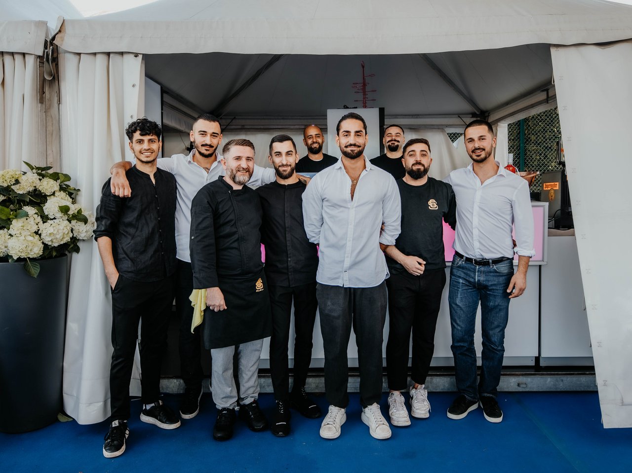 Acht Männer in schwarzer oder weißer Arbeitskleidung stehen in einem Zelt vor einem weißen Vorhang, posieren für ein Gruppenfoto.