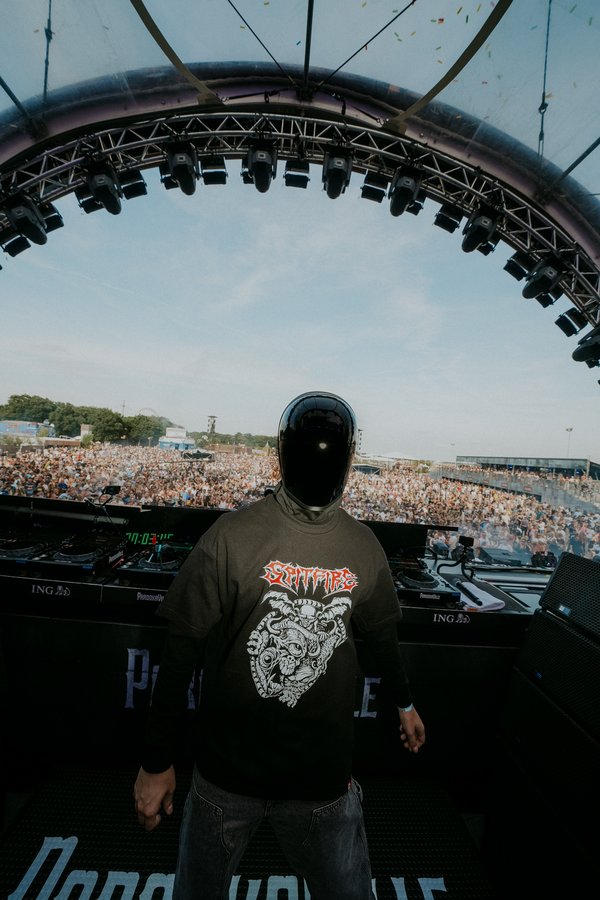Glockenback Eine Person mit einem schwarzen, gesichtsverdeckenden Helm steht auf einer Bühne bei einem Musikfestival. Sie trägt ein dunkles T-Shirt mit einem auffälligen Grafikdruck. Hinter der Bühne erstreckt sich eine große Menschenmenge unter freiem Himmel. Über der Bühne befindet sich eine beeindruckende Lichtkonstruktion. Der blaue Himmel ist im Hintergrund sichtbar.